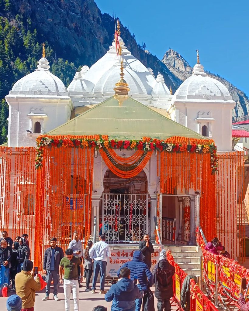 A panoramic view of the Gangotri Temple in Uttarkashi, nestled amidst snow-capped Himalayan peaks. The temple, with its white and pink façade, stands at the base of the towering mountains. The clear blue sky and lush green landscape around the temple highlight its serene and sacred environment