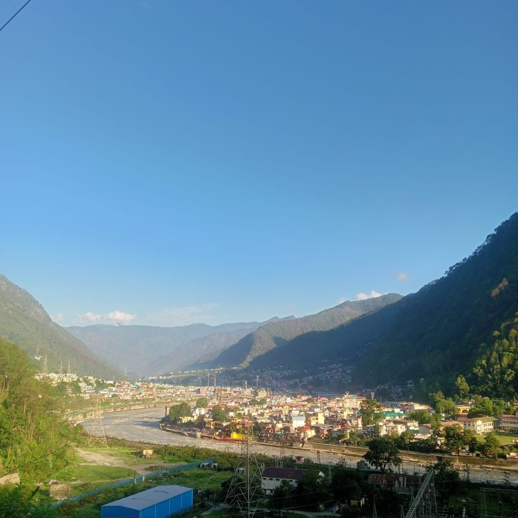 A scenic view of Uttarkashi The serene Bhagirathi River winds through the town, adding to the natural beauty of the landscape. The clear blue sky and surrounding dense forests highlight the town’s tranquil and pristine environment."