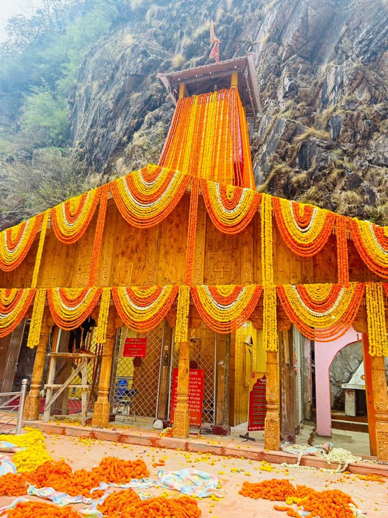 "Yamnotri Temple, nestled in the Himalayas, features a traditional Hindu design with a colorful façade and ornate carvings. The temple is set against a dramatic backdrop of snow-capped peaks and lush, green valleys, creating a serene and majestic atmosphere."